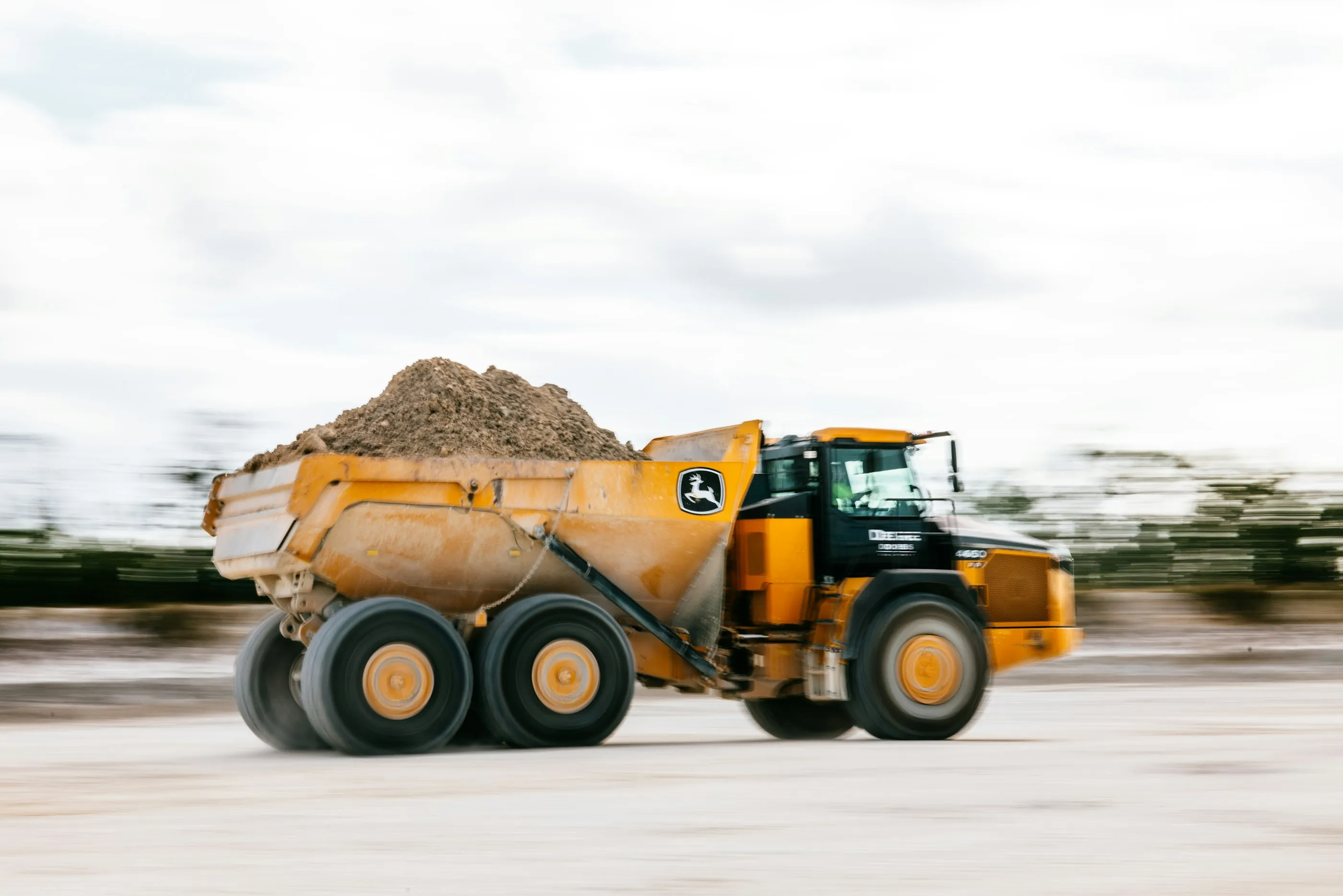 A yellow dump truck carrying a load of soil across a pale, open site