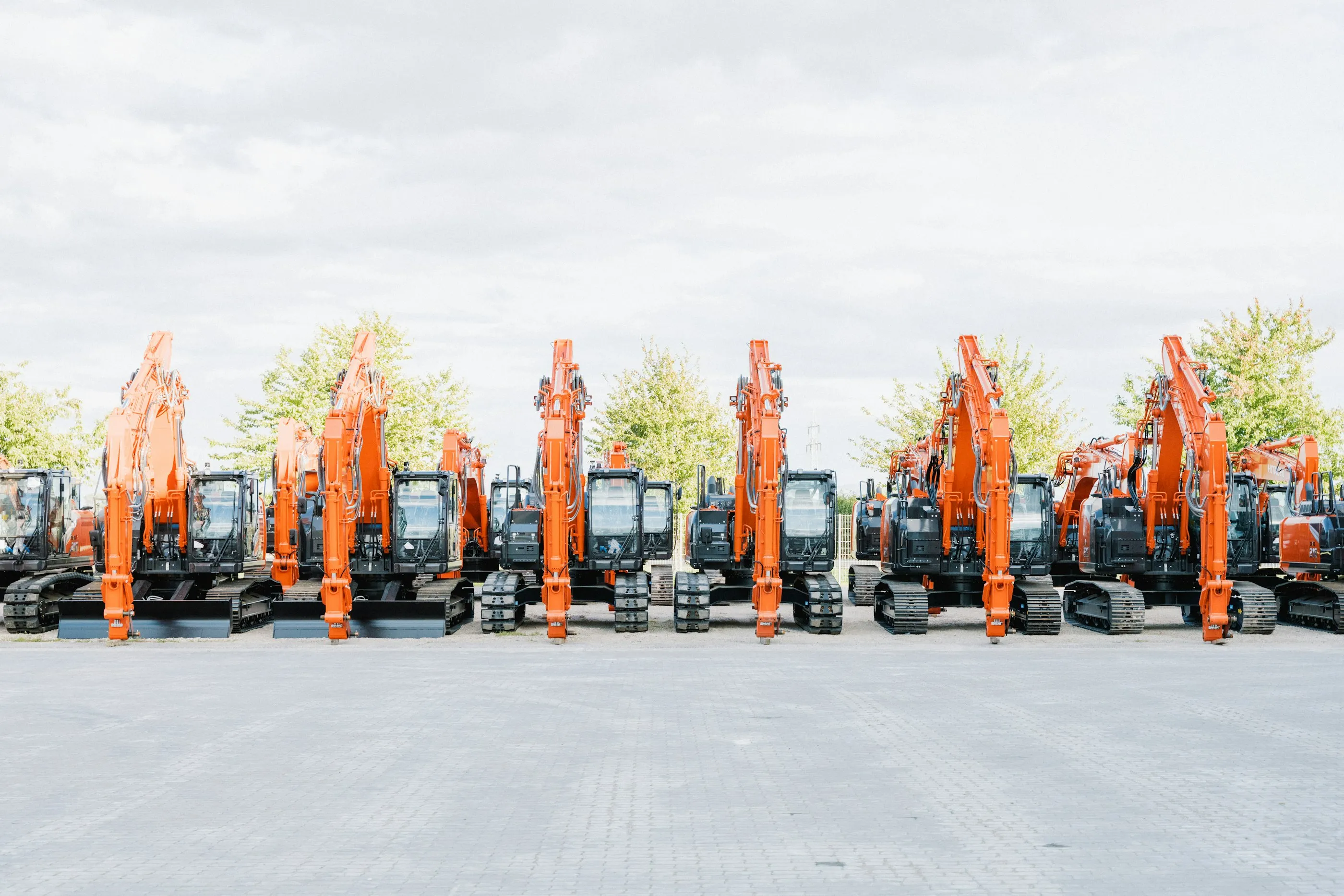 A row of orange excavators parked in a yard, arms raised, under a pale sky