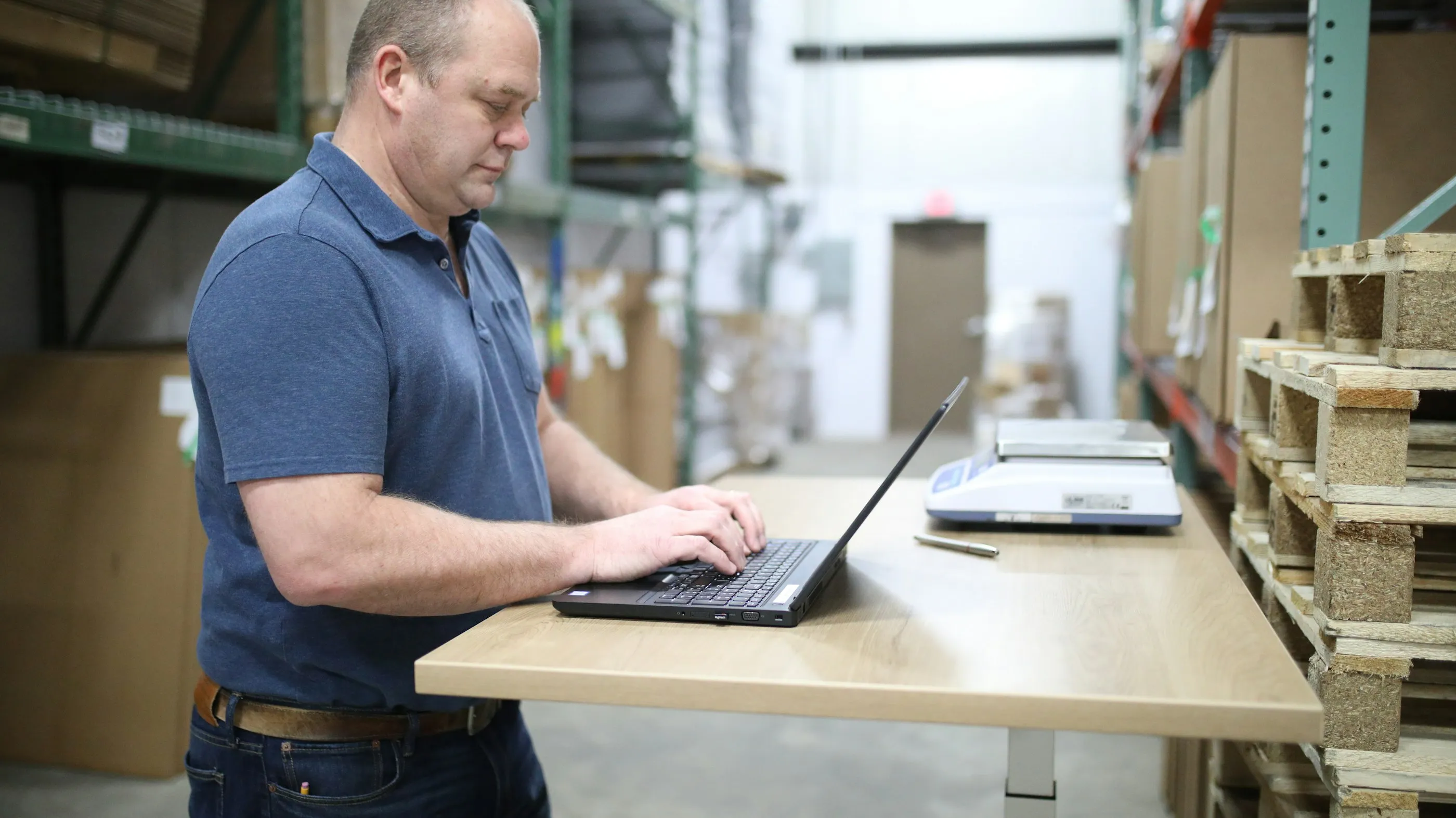 A man working on a laptop at a desk among warehouse shelving and pallets
